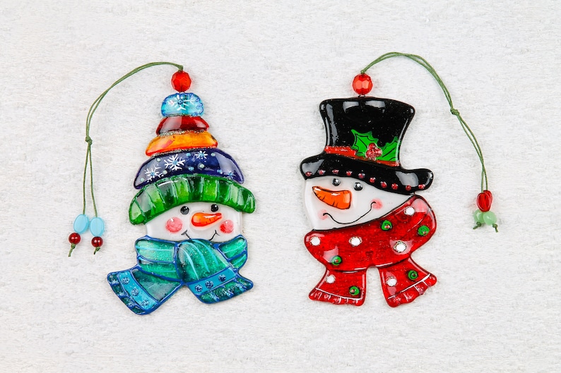 Puede incluir: Dos adornos de vidrio de mu&ntilde;eco de nieve coloridos con detalles verdes, rojos, azules y blancos. El mu&ntilde;eco de nieve de la izquierda tiene un sombrero verde y una bufanda azul. El mu&ntilde;eco de nieve de la derecha tiene un sombrero de copa negro y una bufanda roja.
