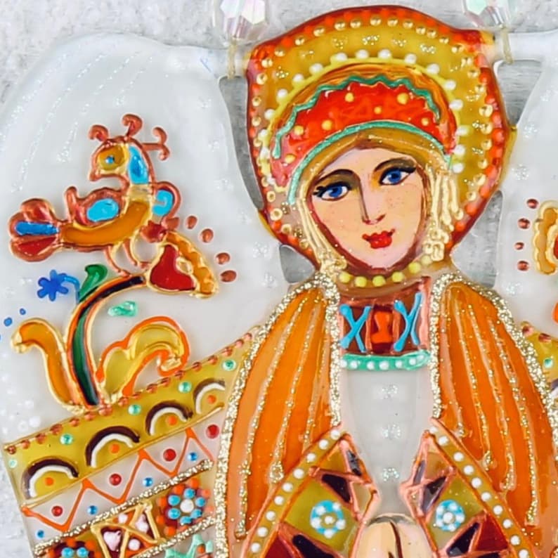 May include: A hand-painted glass angel ornament with a white background and intricate details. The angel is wearing a red and orange dress with gold accents and a crown. The angel has blue eyes and red lips. The ornament is decorated with a bird and floral designs.