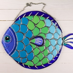 May include: A blue, green, and white glass fish with a silver-colored scale pattern. The fish has a large eye and a small mouth. It is hanging from a silver chain.