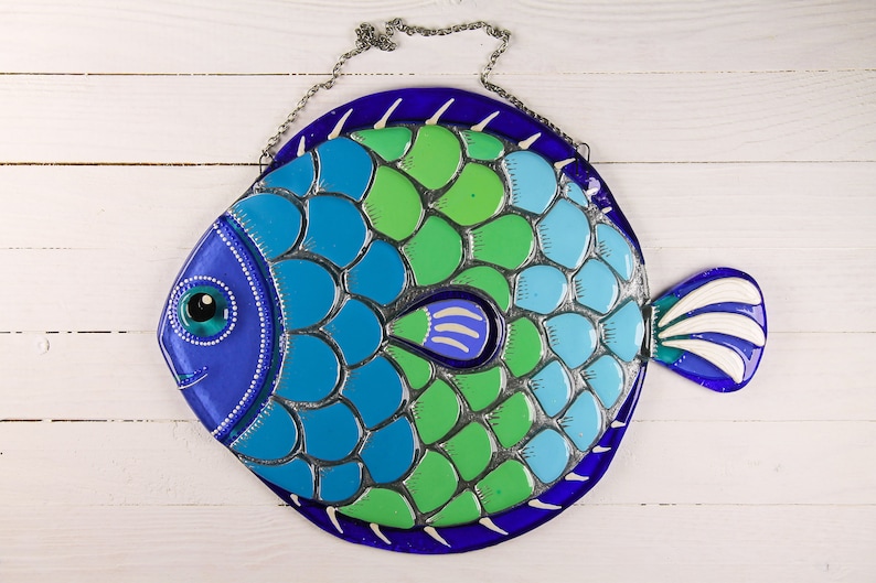 May include: A blue, green, and white glass fish with a silver-colored scale pattern. The fish has a large eye and a small mouth. It is hanging from a silver chain.