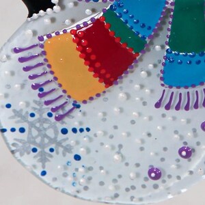 Pode incluir: Enfeite de boneco de neve com um cachecol colorido e detalhes de floco de neve. O cachecol apresenta sec&ccedil;&otilde;es vermelhas, amarelas, verdes e azuis. O enfeite tem uma base branca com detalhes prateados e azuis, e pontos roxos.