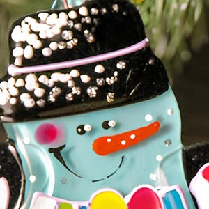 May include: A blue glass snowman ornament with a black hat, white snow, and a red nose. The snowman is wearing a purple scarf and has a white and yellow striped hat.