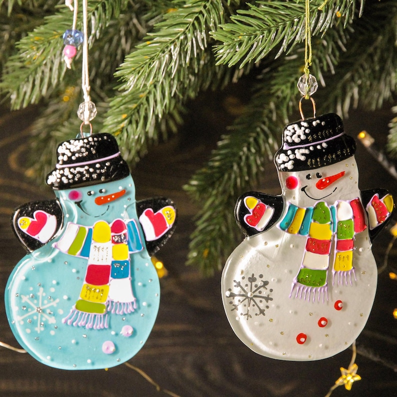 May include: Two glass snowman ornaments with black hats, colorful scarves, and snowflake designs. One snowman is blue and the other is white.