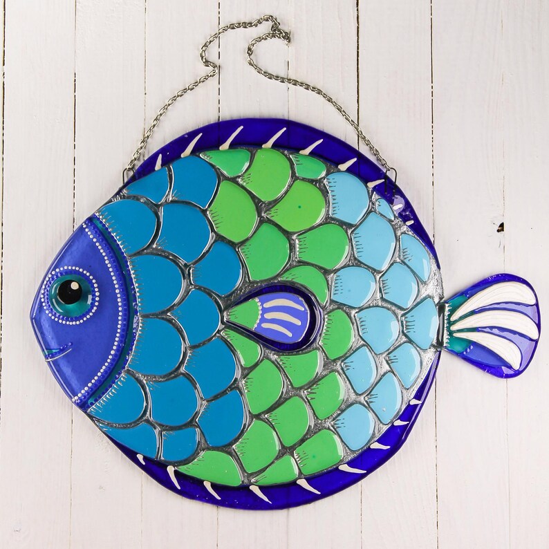 May include: A blue, green, and white stained glass fish with a silver outline. The fish has a blue eye and a white fin. The fish is hanging from a silver chain.