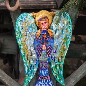 May include: A colorful glass angel ornament with blue, green, and gold wings. The angel is holding a flower in its hands and has a crown on its head.