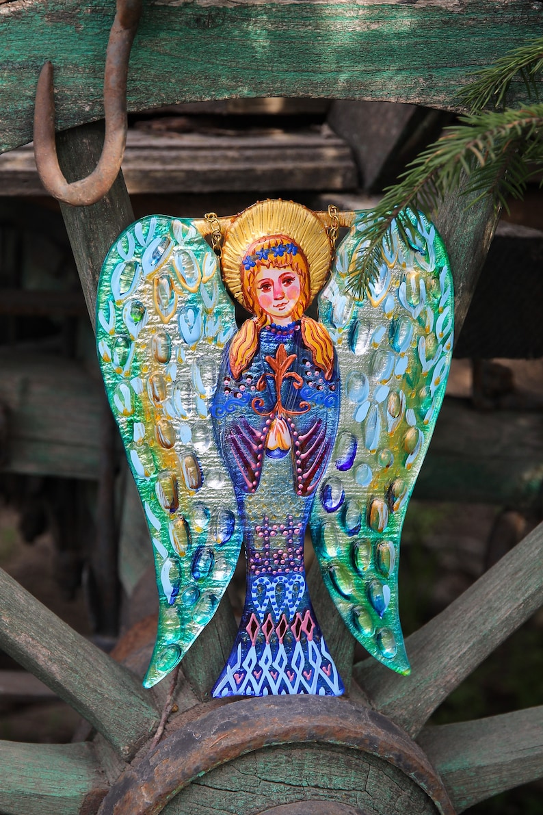May include: A colorful glass angel ornament with blue, green, and gold wings. The angel is holding a flower in its hands and has a crown on its head.