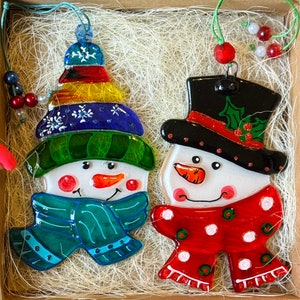 Puede incluir: Dos adornos de vidrio de mu&ntilde;eco de nieve con detalles pintados. Un mu&ntilde;eco de nieve lleva un sombrero azul y verde y una bufanda turquesa. El otro mu&ntilde;eco de nieve lleva un sombrero de copa negro con una banda roja y una bufanda de lunares rojos y blancos.