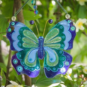 Puede incluir: Una mariposa de vidrio azul, verde y morado con detalles de purpurina cuelga de una rama. La mariposa tiene un dise&ntilde;o detallado con c&iacute;rculos y espirales.