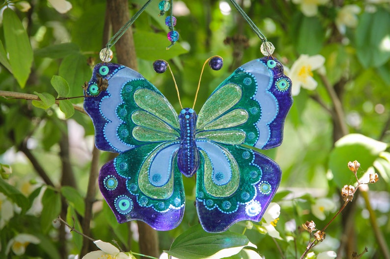 Puede incluir: Una mariposa de vidrio azul, verde y morado con detalles de purpurina cuelga de una rama. La mariposa tiene un dise&ntilde;o detallado con c&iacute;rculos y espirales.