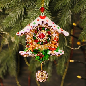Könnte beinhalten: Ein Lebkuchenhaus-Weihnachtsschmuck mit einem Zifferblatt, Lebkuchenmännchen und Stechpalmenakzenten. Der Schmuck ist mit weißem Zuckerguss, roten, grünen und goldenen Details verziert und hat eine kleine Lebkuchenkugel.