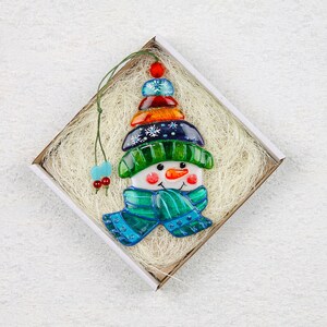 Puede incluir: Un adorno de vidrio de mu&ntilde;eco de nieve con un sombrero azul, verde y naranja y una bufanda azul. El adorno est&aacute; en una caja blanca con papel triturado.
