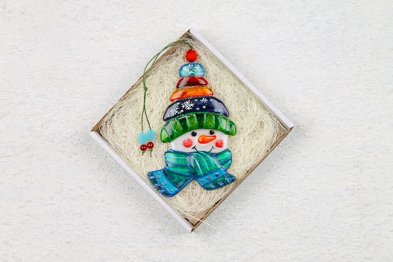 Puede incluir: Un adorno de vidrio de mu&ntilde;eco de nieve con un sombrero azul, verde y naranja y una bufanda azul. El adorno est&aacute; en una caja blanca con papel triturado.