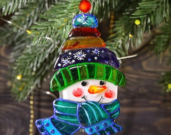 Op de afbeelding: Een sneeuwpopornament van gefuseerd glas met een veelkleurige hoed met sneeuwvlokdetails, een groene geribbelde rand, een wit gezicht met een wortelneus en een blauwe sjaal, opgehangen aan een groen koord met een rode kraal.