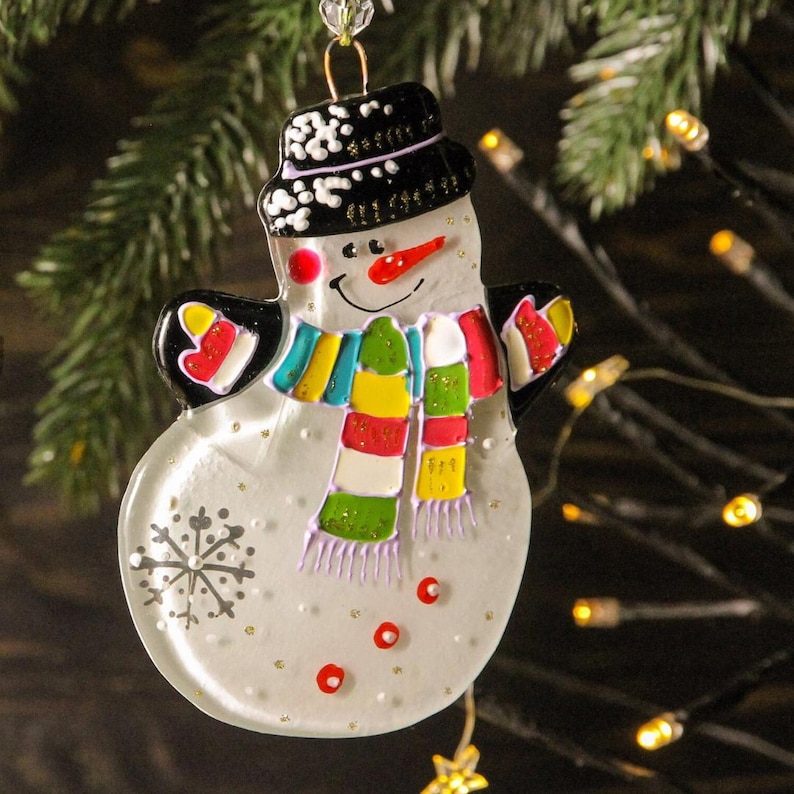Pode incluir: Enfeite de Natal de boneco de neve com cartola preta, cachecol colorido e nariz de cenoura. O enfeite tem um rosto sorridente e &eacute; decorado com um floco de neve e pontos vermelhos. Est&aacute; pendurado em frente a uma &aacute;rvore de Natal.