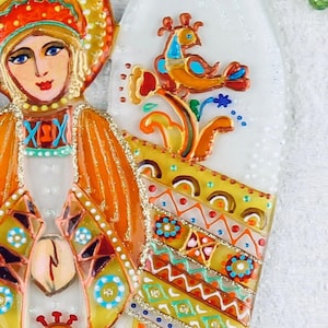 May include: A hand-painted glass ornament featuring a stylized angel with a colorful, patterned dress and a bird perched on a branch. The angel has a halo and a crown, and the ornament is decorated with gold glitter.