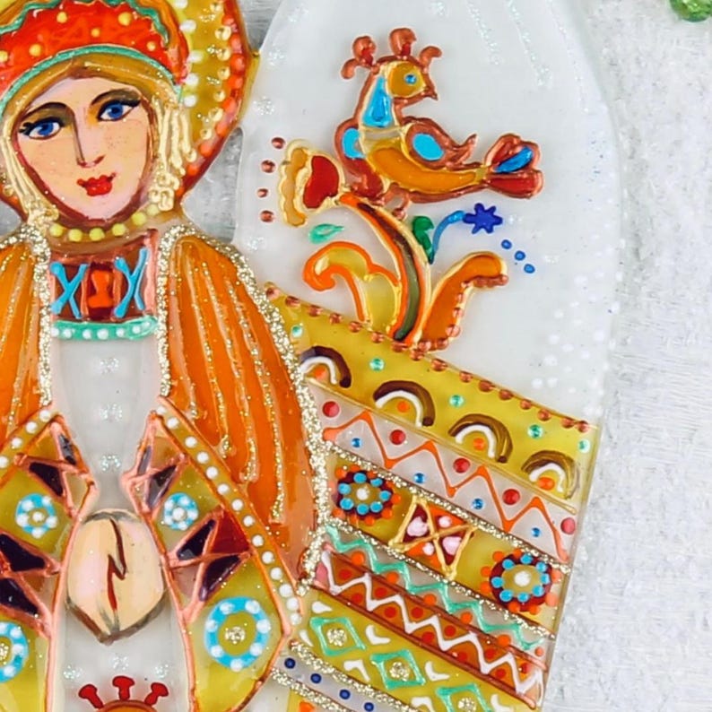 May include: A hand-painted glass ornament featuring a stylized angel with a colorful, patterned dress and a bird perched on a branch. The angel has a halo and a crown, and the ornament is decorated with gold glitter.