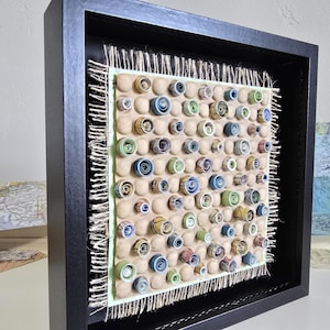 May include: Abstract art piece in a black frame. The artwork features numerous rolled paper elements in various colors, including blue, green, and beige, arranged on a textured background. The frame has a natural fiber border.