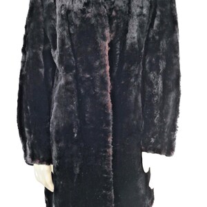 May include: A long, black faux fur coat with a notched collar. The coat has a dark brown trim and a classic, tailored design. The coat is displayed on a mannequin against a white background.
