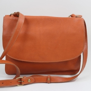 May include: A leather crossbody bag in a warm orange hue. The bag features a flap closure and an adjustable shoulder strap. The bag has a textured surface and a brass buckle. The bag is a stylish accessory.
