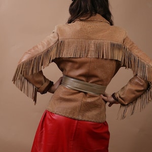 May include: A brown leather jacket with fringe detailing and a matching brown leather belt. The jacket has a long, flowing silhouette and is worn over a red leather skirt.