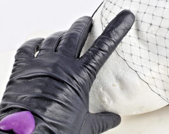 May include: A black leather glove with a purple heart-shaped embellishment. The glove is touching a white object covered with a black net. The glove is made of a smooth, dark material.