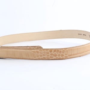May include: Tan leather belt with a crocodile-like embossed pattern. The belt is a light tan color and has a classic design. The belt is made of leather and has a buckle closure. The belt is a versatile accessory for any wardrobe.