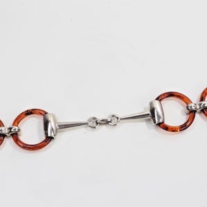 May include: A horse bit with a silver metal and tortoiseshell-patterned rings. The bit is designed with two sets of rings connected by a central chain. The rings are a warm brown color, and the metal has a shiny finish.