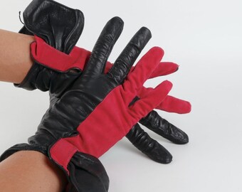 May include: A pair of gloves featuring a color-blocked design with black leather on the outer fingers and palm side, contrasted by vibrant red suede panels on the inner fingers and back of the hand, finished with a gathered black leather cuff.