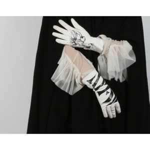 May include: A pair of white gloves with black designs. One glove has a floral design and the other has a geometric design. The gloves are layered over a sheer white fabric with ruffles.