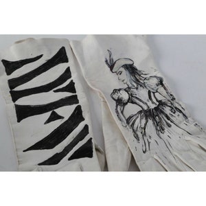 May include: A pair of white leather gloves with hand-painted designs. One glove features a black and white zebra-like pattern, while the other glove has a black and white illustration of a woman in a dress and hat.