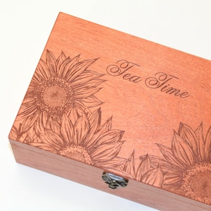 May include: A rectangular wooden box with a light brown finish featuring laser-engraved sunflower illustrations on the lid. The text "Tea Time" is engraved in a cursive script on the top surface. A small metal latch is attached to the front edge.