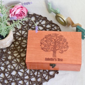 May include: A rectangular wooden box with a hinged lid featuring a laser-engraved tree design with heart-shaped leaves and the text Adelle's Tea. The box has a small metal latch on the front and a smooth, light brown finish.