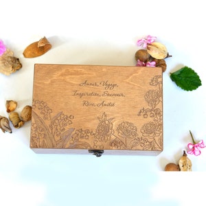 Könnte beinhalten: Eine Holzkiste mit einem Scharnierdeckel und einem gravierten Blumendesign auf der Oberseite. Die Kiste ist geschlossen und der Deckel ist leicht geöffnet. Die Gravur enthält die Worte "Amour, Voyage, Inspiration, Souvenir, Reve, Amitie".