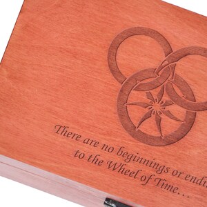 May include: A rectangular wooden box with a reddish-brown finish. The lid features a laser-engraved design of three interlocking rings surrounding a central star-like shape. Below the design, the text reads: There are no beginnings or endings to the Wheel of Time...