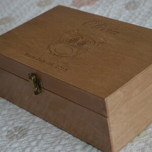 May include: A wooden box with a hinged lid and a brass latch. The box is engraved with the name "Olivia" and the date "Born July 25, 2018". The box also features a carved owl design.