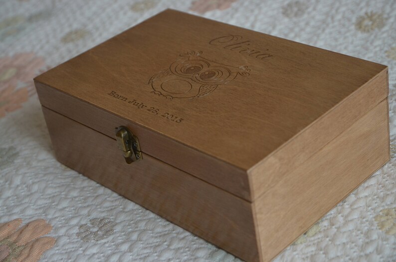 May include: A wooden box with a hinged lid and a brass latch. The box is engraved with the name "Olivia" and the date "Born July 25, 2018". The box also features a carved owl design.