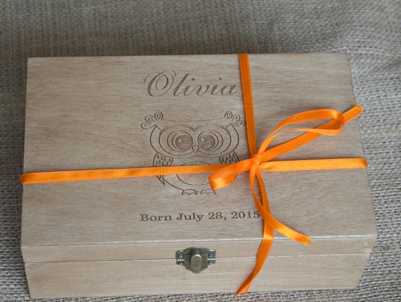 May include: A wooden box with an orange ribbon tied around it. The box is engraved with the name "Olivia" and an owl design. The text "Born July 28, 2015" is also engraved on the box.