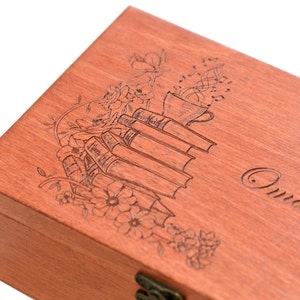 May include: A rectangular wooden box with a reddish-brown finish featuring a laser-engraved design on the lid. The engraving depicts a stack of books, a steaming cup, and floral elements, with the text "Oma's" visible on the right side.