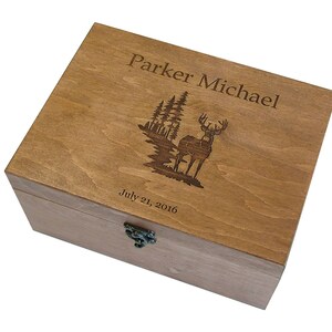 Op de afbeelding: Een houten doos met een scharnierend deksel en een metalen sluiting. Het deksel is gegraveerd met een herten silhouet, bomen en de tekst "Parker Michael July 21, 2016".