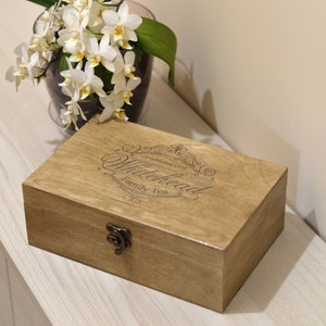 May include: A rectangular wooden box with a hinged lid and a metal latch. The top of the box features an engraved design with the text "Whitehead Family Tea" and "Established 1939" surrounded by decorative flourishes in a dark brown finish.