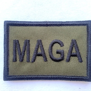 May include: An olive green rectangular patch with the word "MAGA" embroidered in black block letters. The patch has a black border.