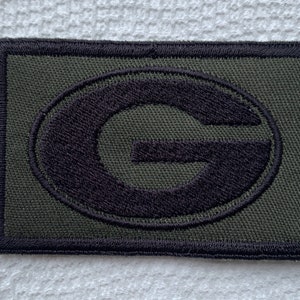May include: A rectangular patch with a dark green background and a black embroidered "G" logo. The logo is centered within the patch, and the edges are outlined with black stitching. The patch is likely for apparel or accessories.