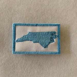 May include: Embroidered patch featuring the outline of North Carolina in light blue. The patch has a white background and a light blue border. The patch is rectangular in shape and appears to be made of fabric.