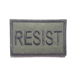 May include: An olive green rectangular patch with the word "RESIST" embroidered in black. The patch has a black border and is set against a white background. The text is in a bold, sans-serif font.