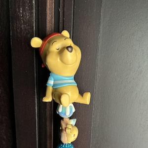 May include: A yellow Winnie the Pooh figurine wearing a red headband and a light blue and white striped shirt, sitting on a small blue and white sphere, with a smaller tan character figure attached below it on a blue beaded base.