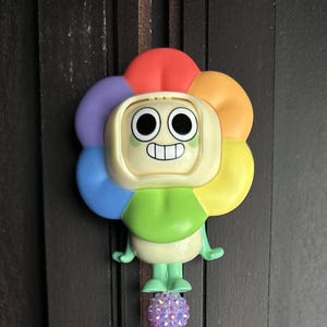 May include: A decorative wall ornament featuring a cartoon character with a large, multi-colored flower-shaped head. The petals are red, orange, yellow, green, blue, and purple. The face is cream-colored with large black eyes and a wide, toothy grin.