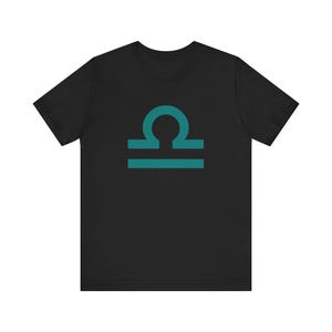 May include: Black t-shirt featuring a teal Libra zodiac symbol. The graphic is centered on the front of the shirt. The shirt has short sleeves and a crew neck.