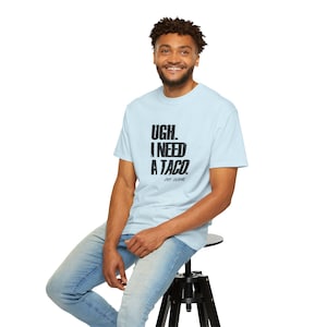 Puede incluir: Una camiseta azul claro con el texto "UGH. I NEED A TACO." en negro. La camiseta la lleva una persona sentada en un taburete negro, con vaqueros azules. El texto "JOY DOME" está debajo.