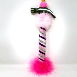 May include: A decorative hammer with a silver head and a handle wrapped in pink, purple, and silver ribbon. The handle is adorned with pink feathers and a pink pom-pom. A small gold-colored pom-pom sits atop the hammer head.