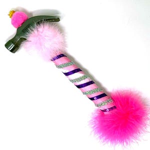 May include: A decorative hammer with a green head and a pink, embellished handle. The handle is wrapped with pink and purple ribbons, sequins, and feathers. A pink pom-pom and a small gold crown adorn the hammer head.